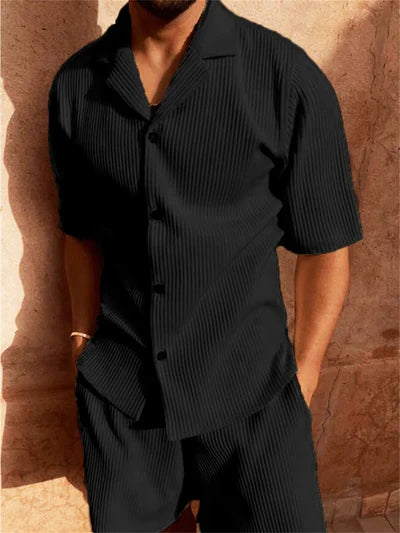 Person wearing a black pleated shirt and pants against a textured wall.